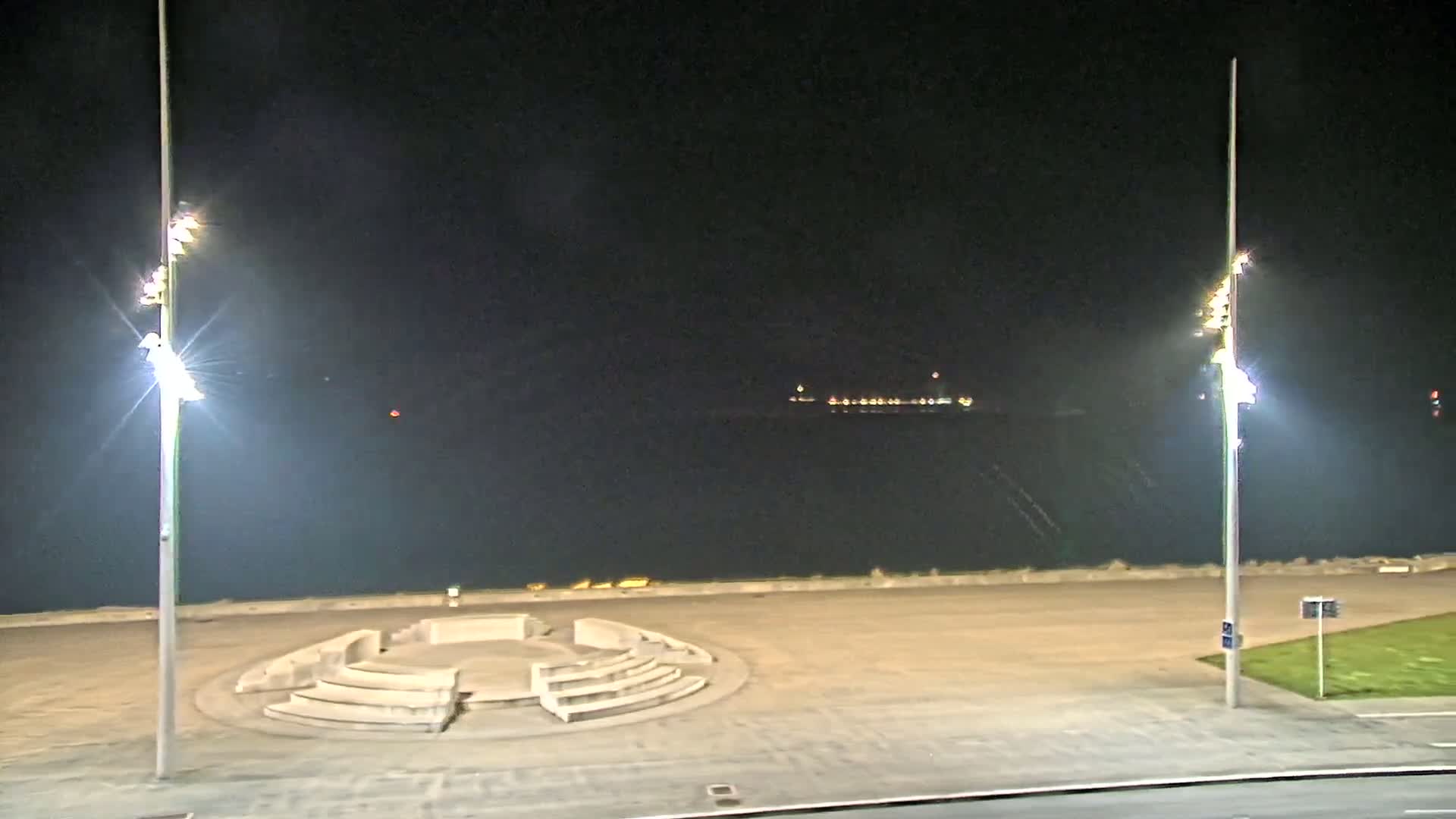 A nighttime view of a paved waterfront area with a circular seating structure, two tall illuminated lamp posts, and a dark body of water with distant lights in the background.