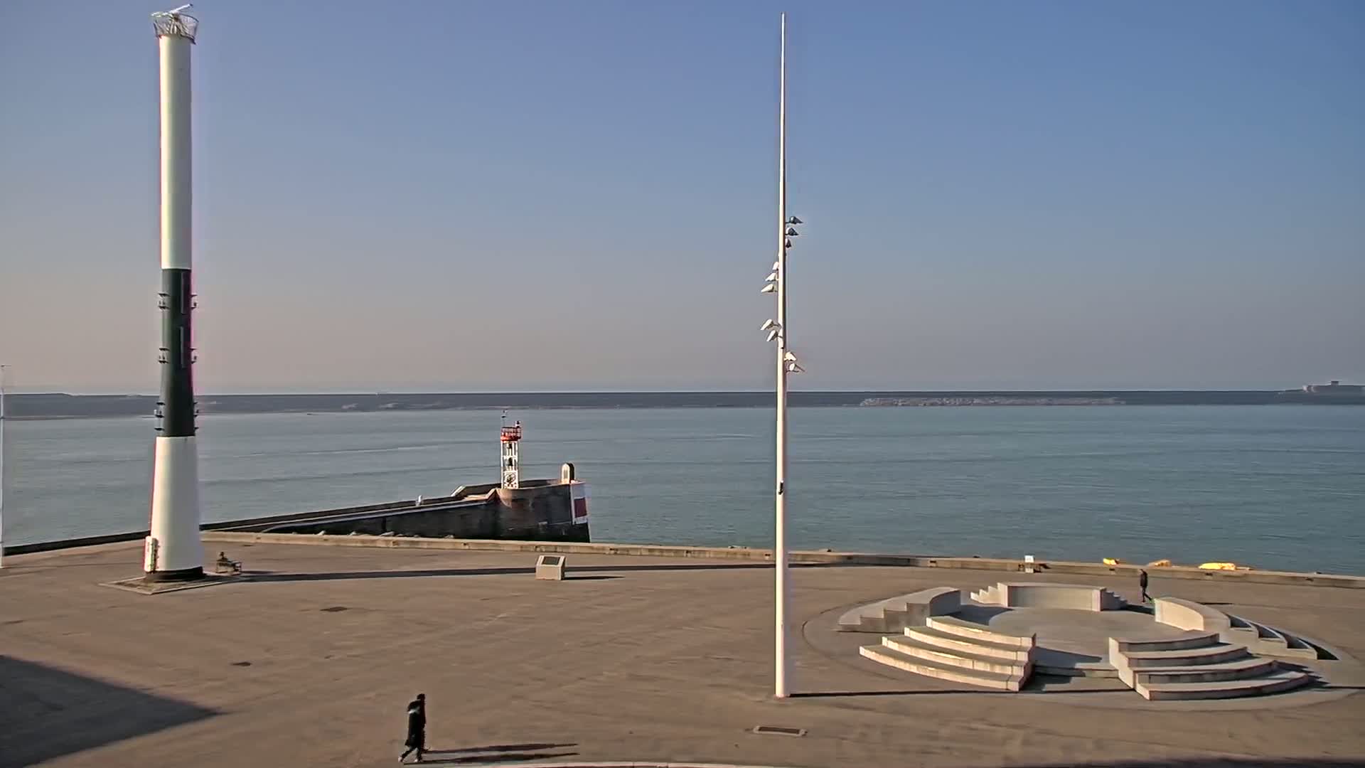 A tall, black and white lighthouse stands near a concrete amphitheater overlooking a calm body of water under a clear, sunny sky.