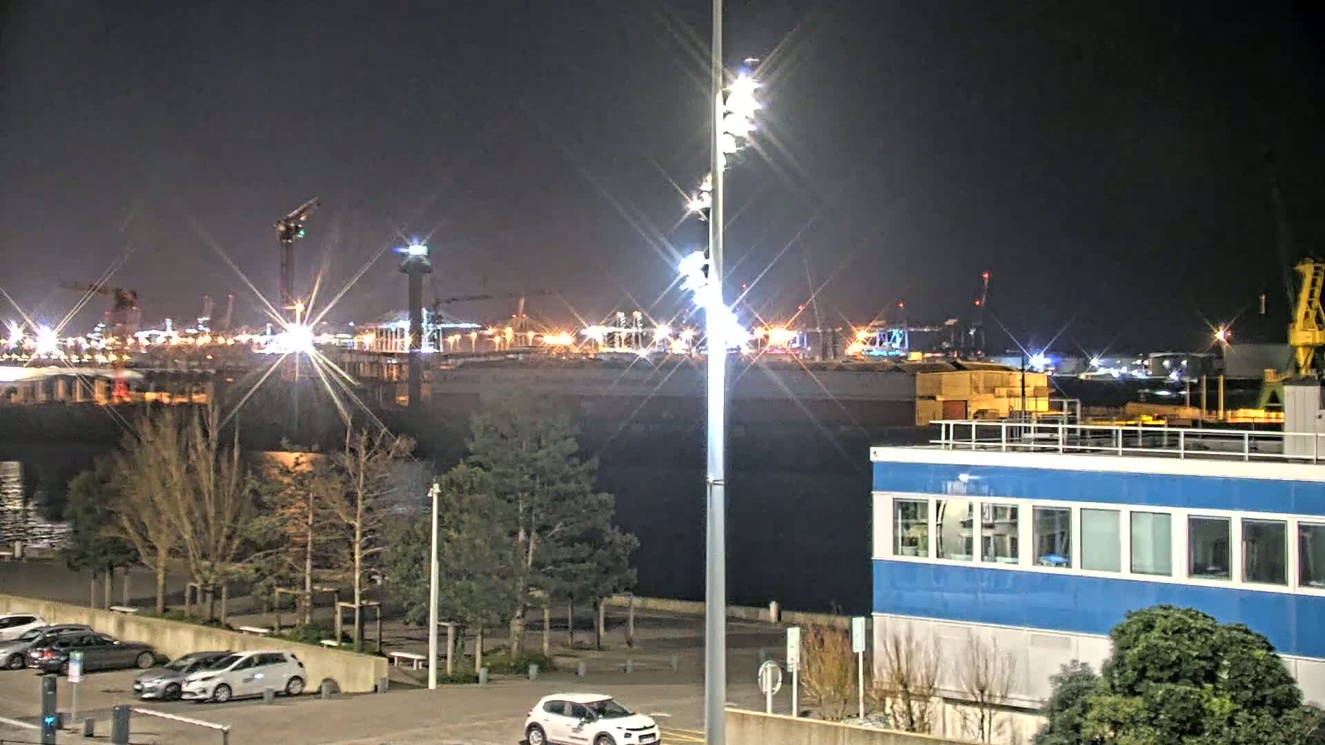 A brightly lit industrial waterfront at night, featuring several cranes and a large blue building, with cars parked in a lot in the foreground.
