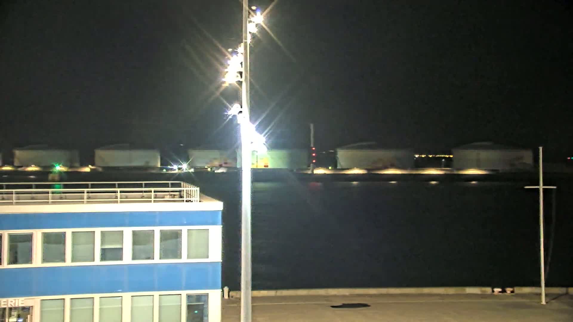 At night, a brightly lit tall pole stands near a blue building overlooking a dark body of water with several illuminated storage tanks in the background.
