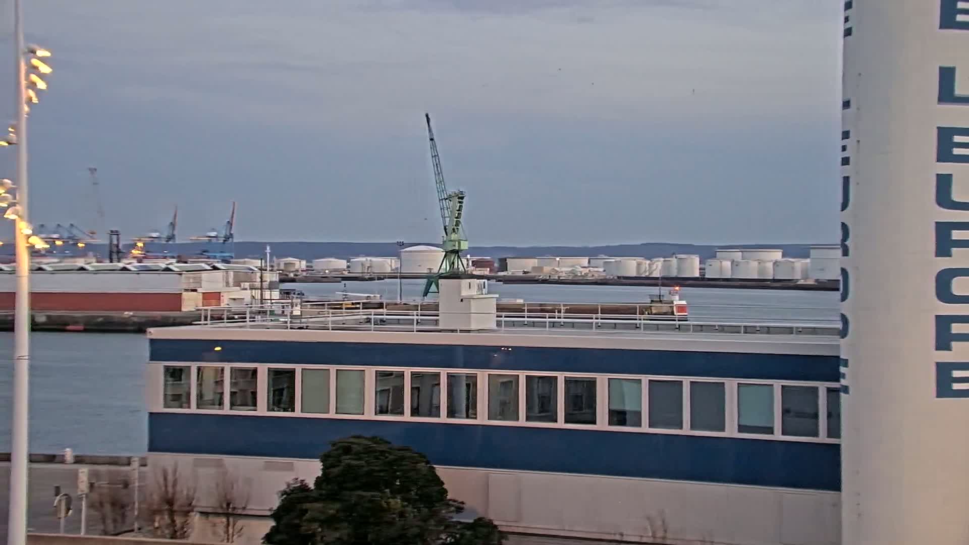 A blue building with many windows sits on a waterfront, a crane stands on its roof, and numerous storage tanks and industrial structures are visible in the background under an overcast sky.