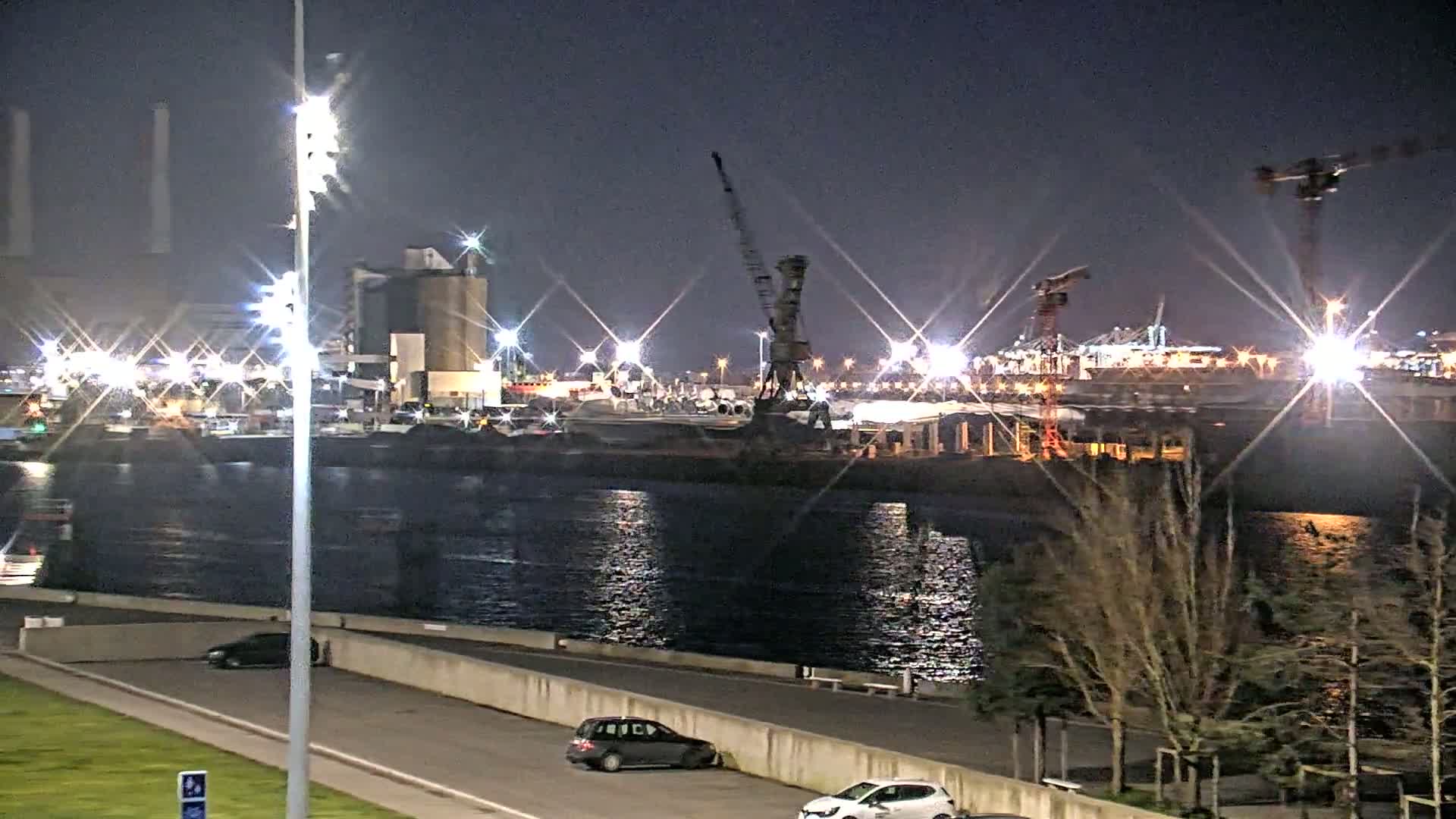 A nighttime view of a harbor with industrial buildings, cranes, and several cars parked along a waterfront road.