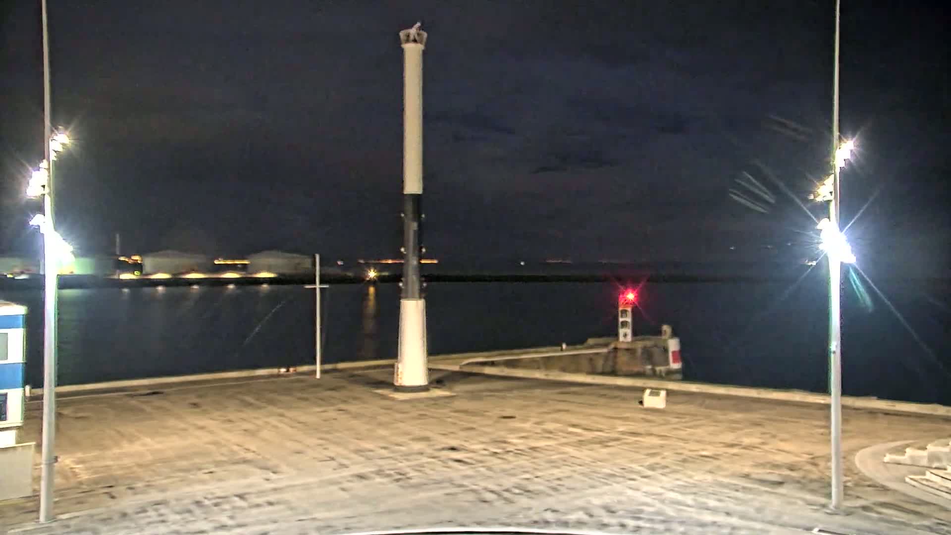 A tall, slender lighthouse stands in a dark, empty harbor at night, illuminated by bright lights on either side, with calm water and distant lights visible across the bay.
