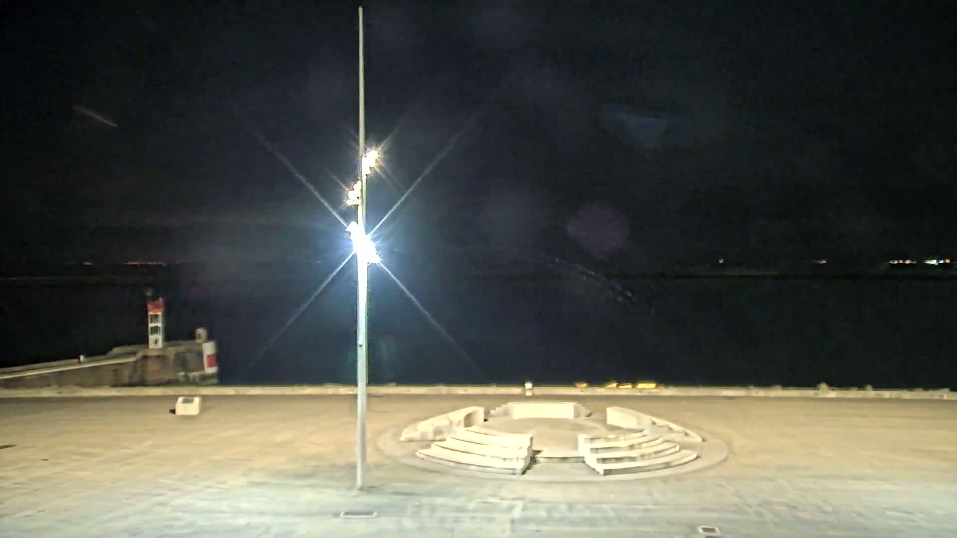 A brightly lit, tall lamppost stands in a nighttime seaside plaza featuring a circular stone seating area, with a calm, dark body of water visible in the background.