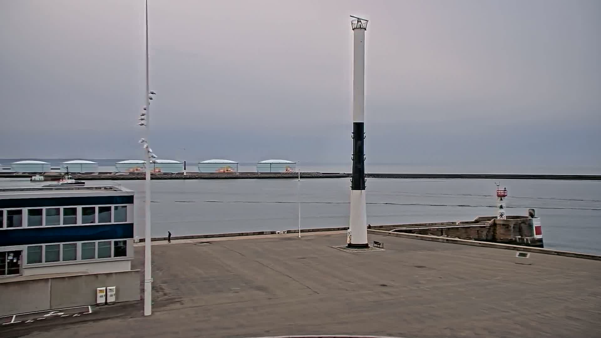 A tall, slender lighthouse stands in a harbor on an overcast day, with several storage tanks visible in the distance and a small building to the left.