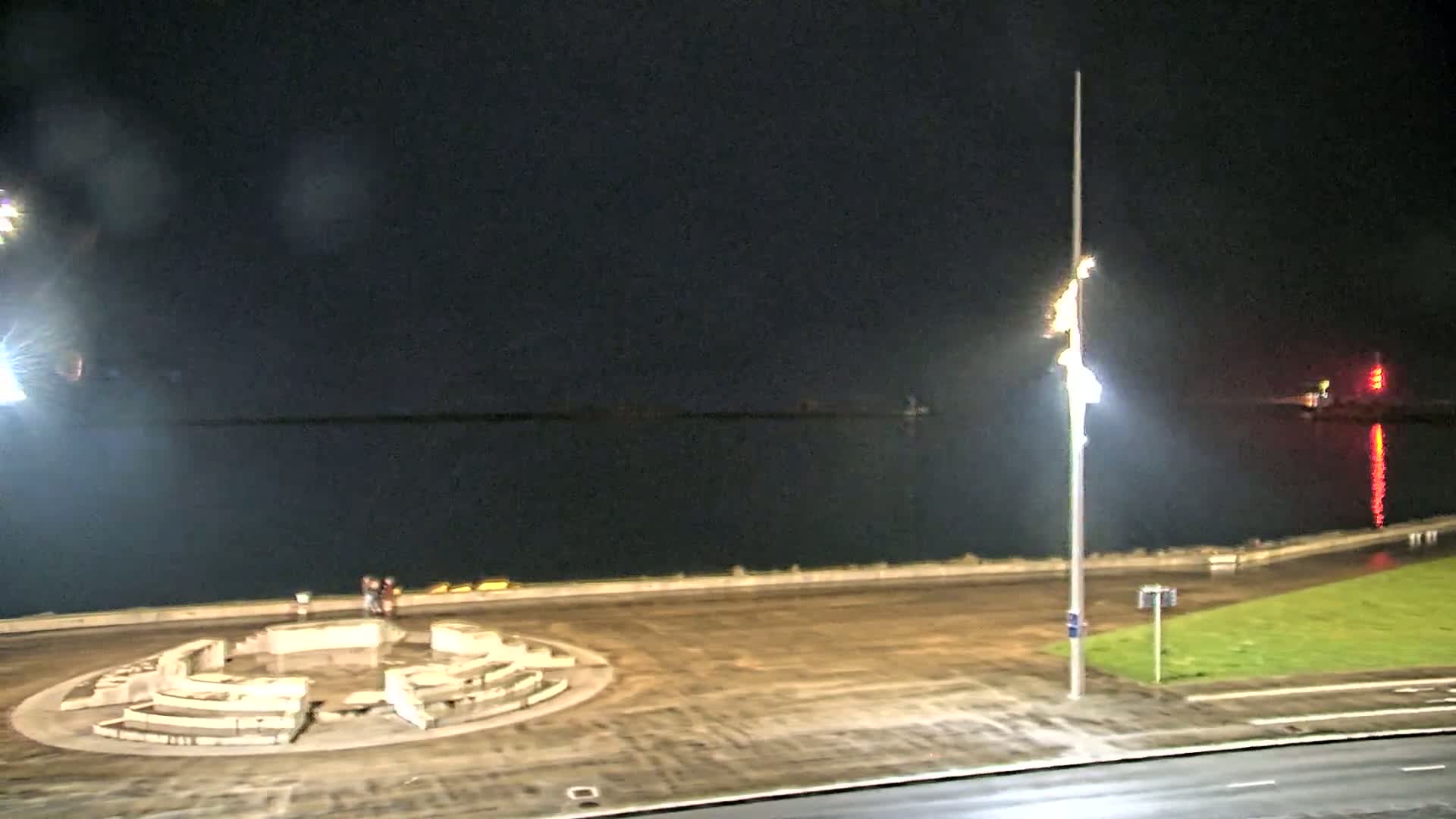 A nighttime view of a calm body of water, a paved area with a circular stone structure, and a roadway, illuminated by artificial lights under a dark sky.