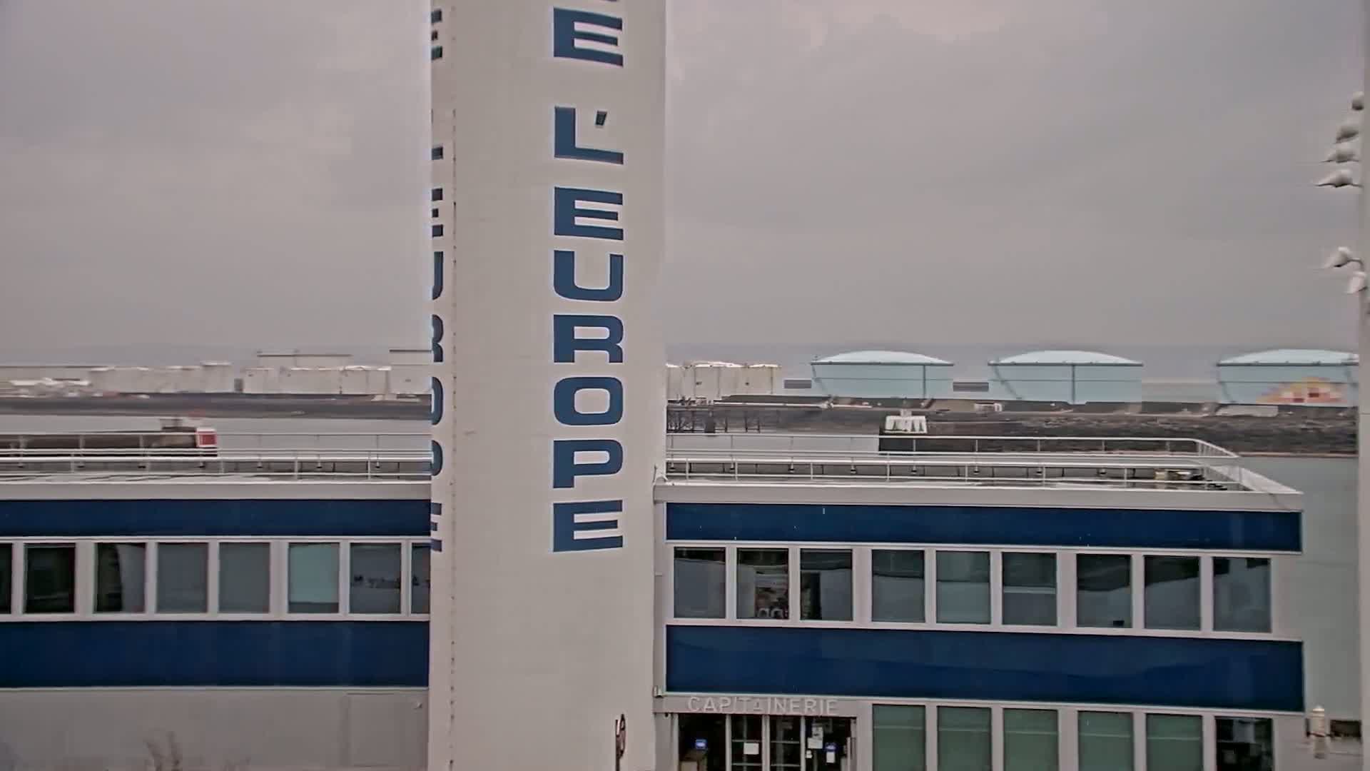 A tall white column with "L'EUROPE" written on it stands next to a blue-trimmed building, overlooking a body of water with several large, light-blue storage tanks in the distance under an overcast sky.