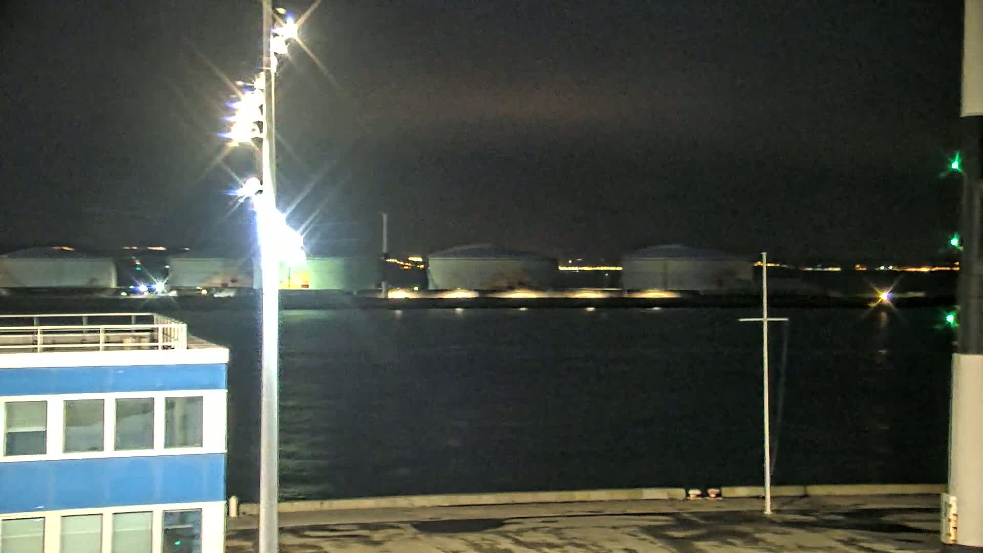 At night, across a dark body of water, several large storage tanks are illuminated, a bright light post stands near a two-story blue building.