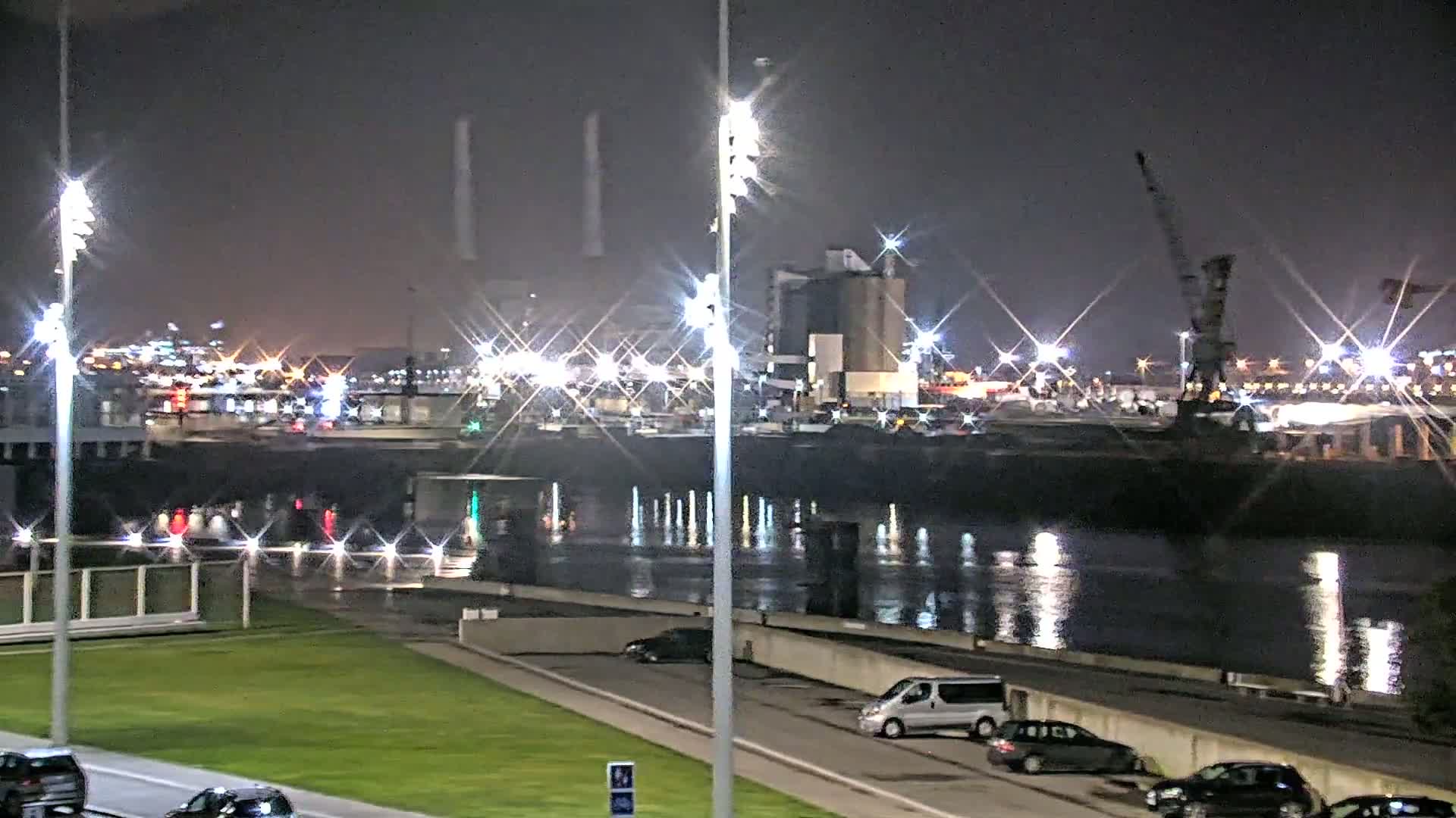 A nighttime view of a calm body of water reflecting the lights of a distant industrial area, with several cars parked along a grassy waterfront.