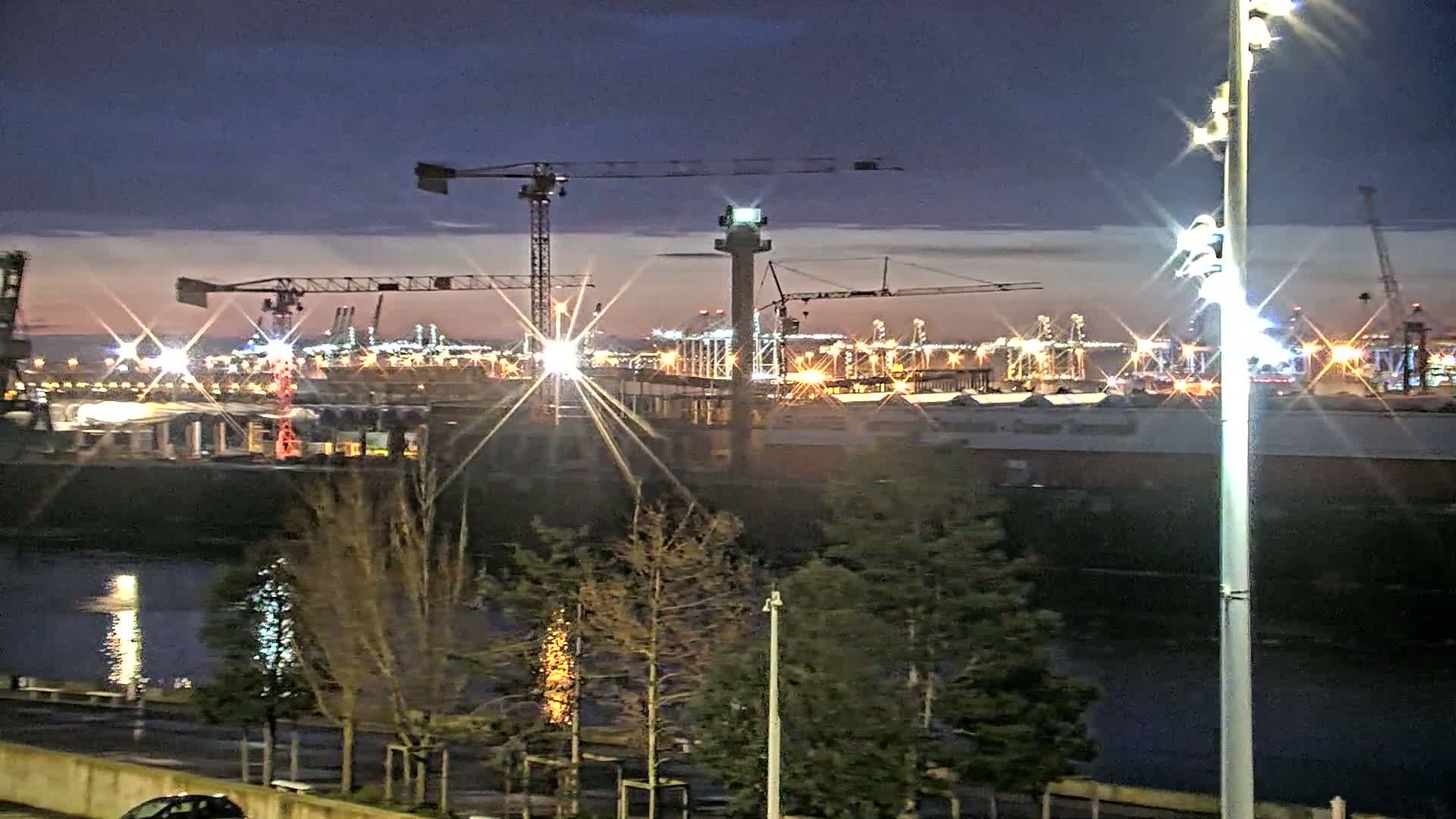 A brightly lit industrial port at night is seen across a body of water, with several cranes and other construction equipment visible, under a twilight sky.