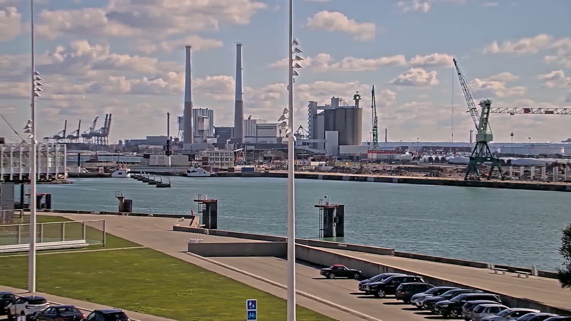 A partly cloudy day shows a harbor with industrial buildings, cranes, and several docked boats, a grassy area with parked cars, and calm water.