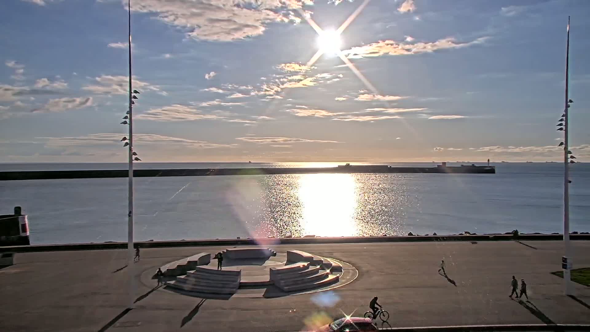 A sunlit seaside promenade with a circular amphitheater-like structure, a few pedestrians, and a cyclist, under a partly cloudy sky.