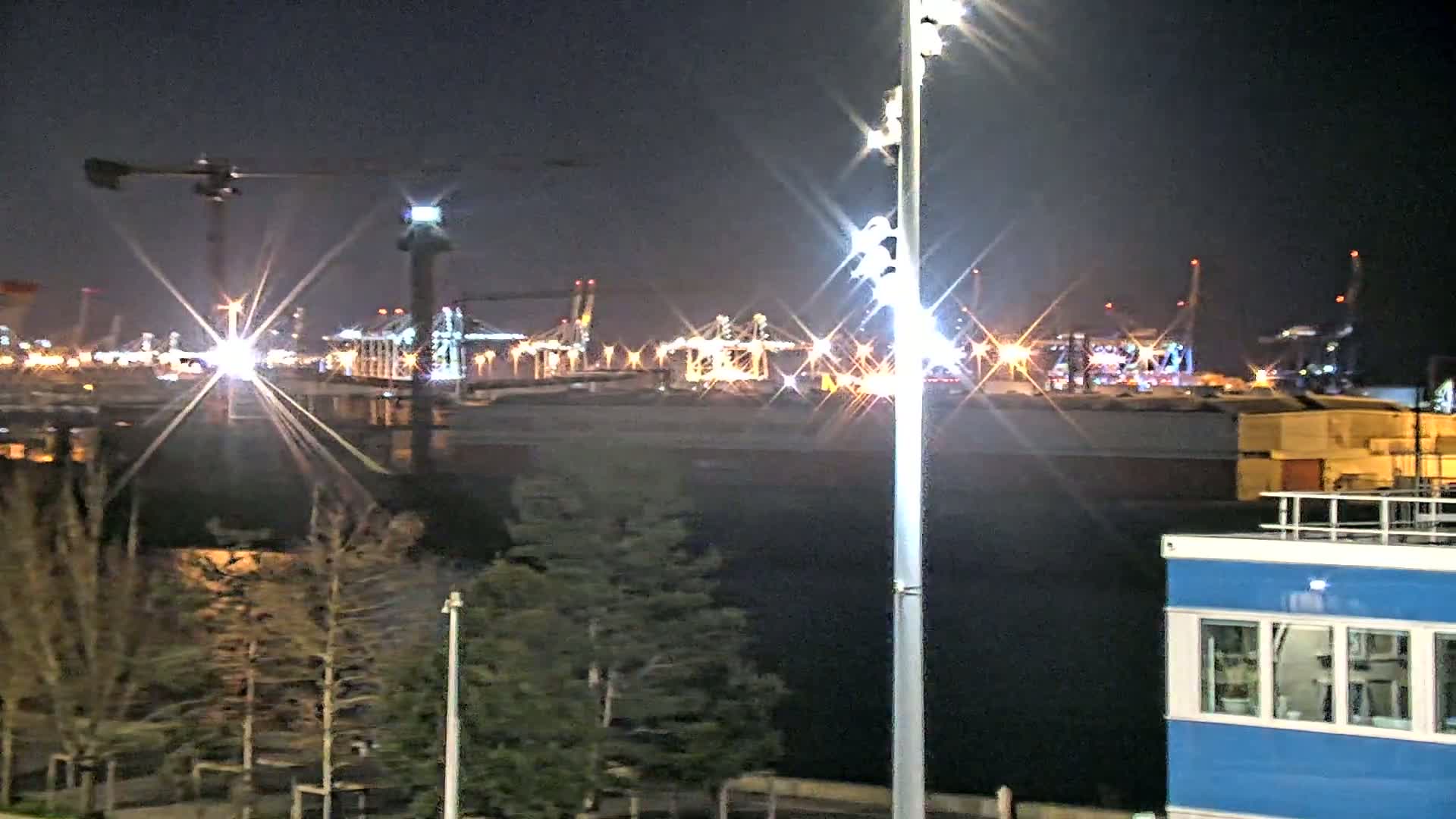 A brightly lit industrial port at night, viewed from across a body of water, with trees and a small blue building in the foreground.