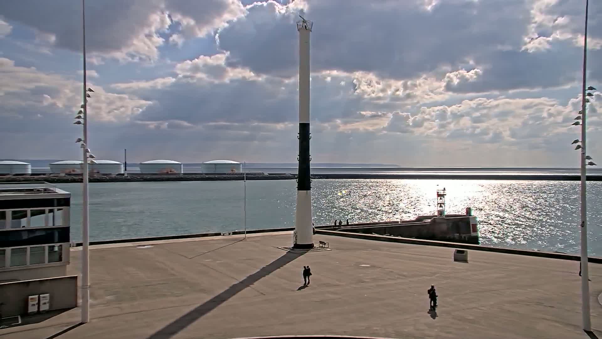 A tall, slender lighthouse stands in a harbor area under a partly cloudy sky, with a calm body of water reflecting sunlight and several large storage tanks visible in the distance.