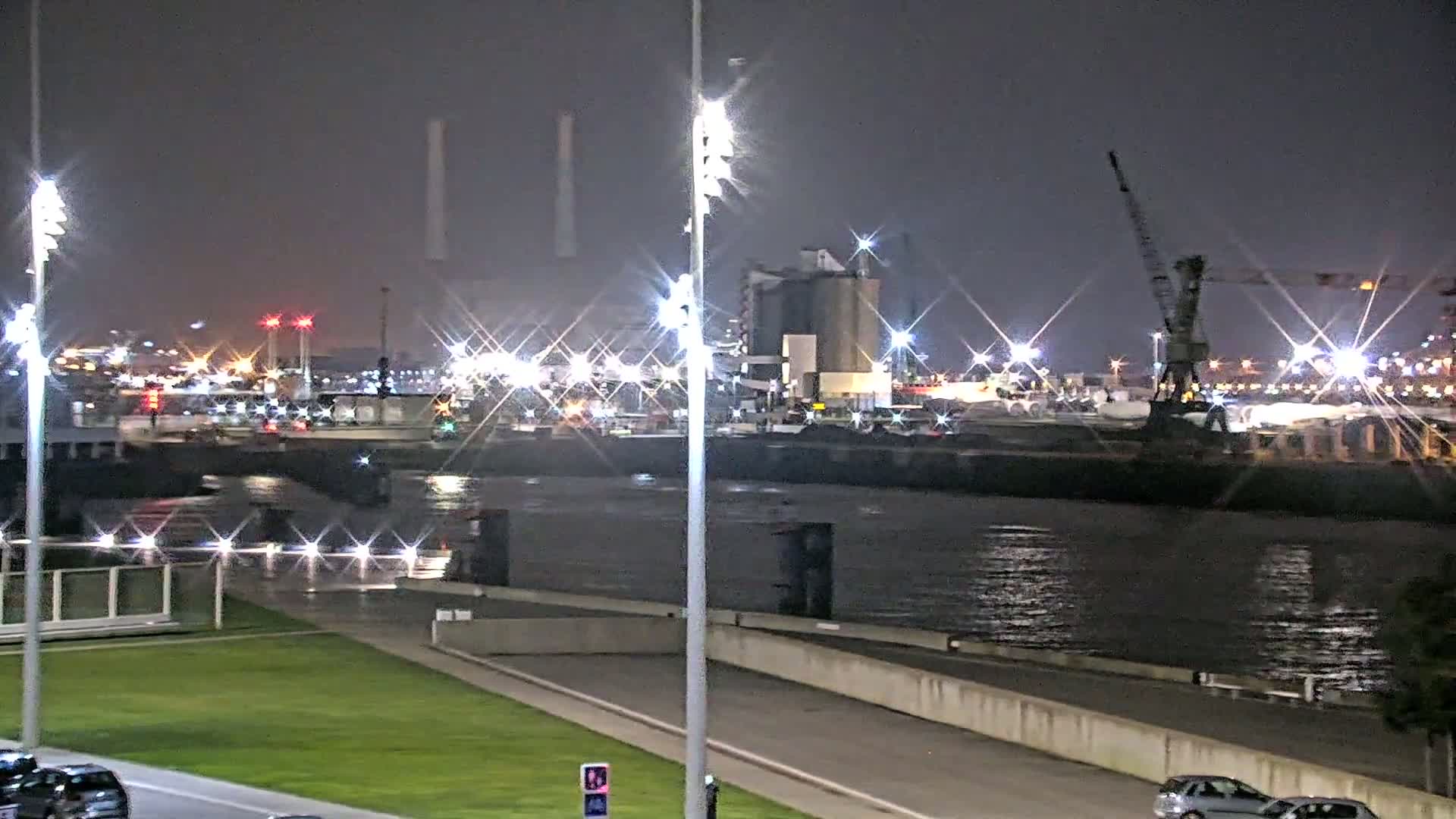 A nighttime view of a harbor with brightly lit industrial buildings and cranes along a calm body of water, with a grassy area and roadway in the foreground.