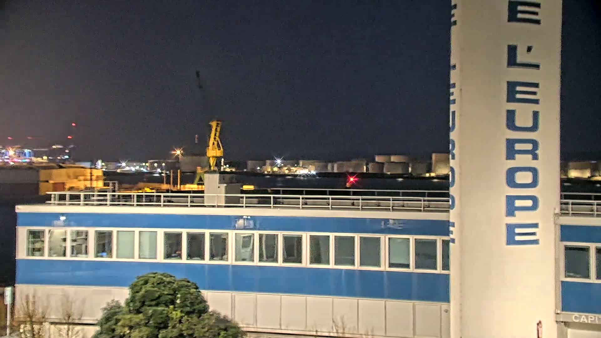 At night, a blue building with many windows sits in front of a body of water, beyond which are industrial structures and a yellow crane.