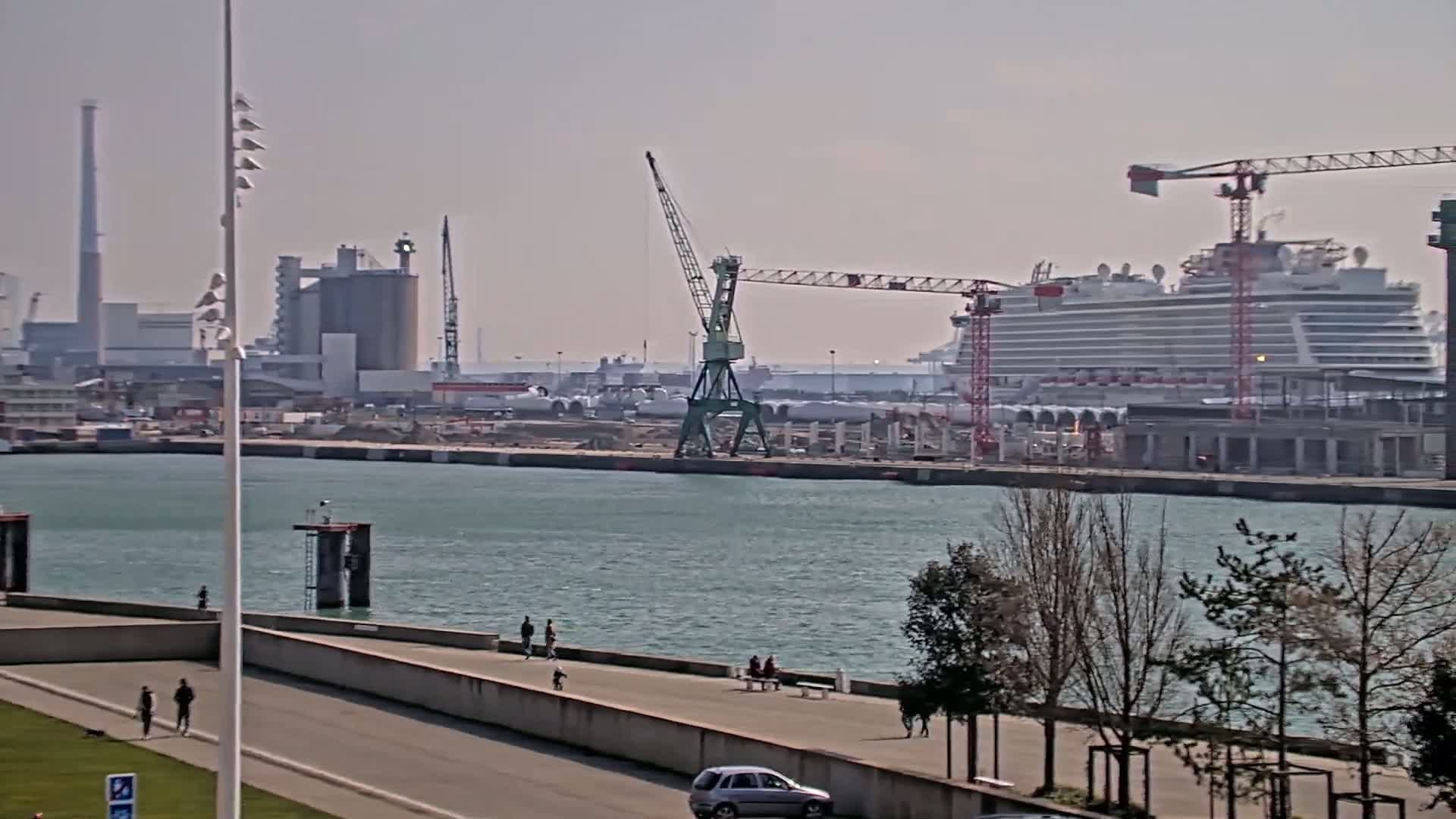 A large cruise ship is docked at a shipyard under a hazy sky, with several cranes and industrial buildings in the background, and people walking along a waterfront path in the foreground.