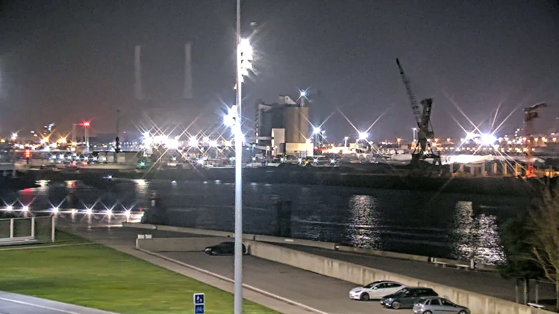 A nighttime view of a harbor, with brightly lit industrial buildings and cranes across a body of water from a grassy area with parked cars.