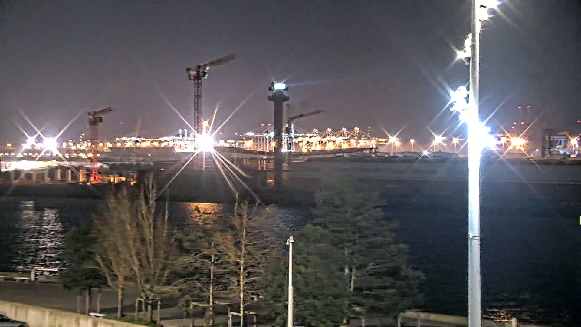 A nighttime view of a harbor with several cranes, a large ship, and brightly lit industrial structures across the water, under a clear, dark sky.