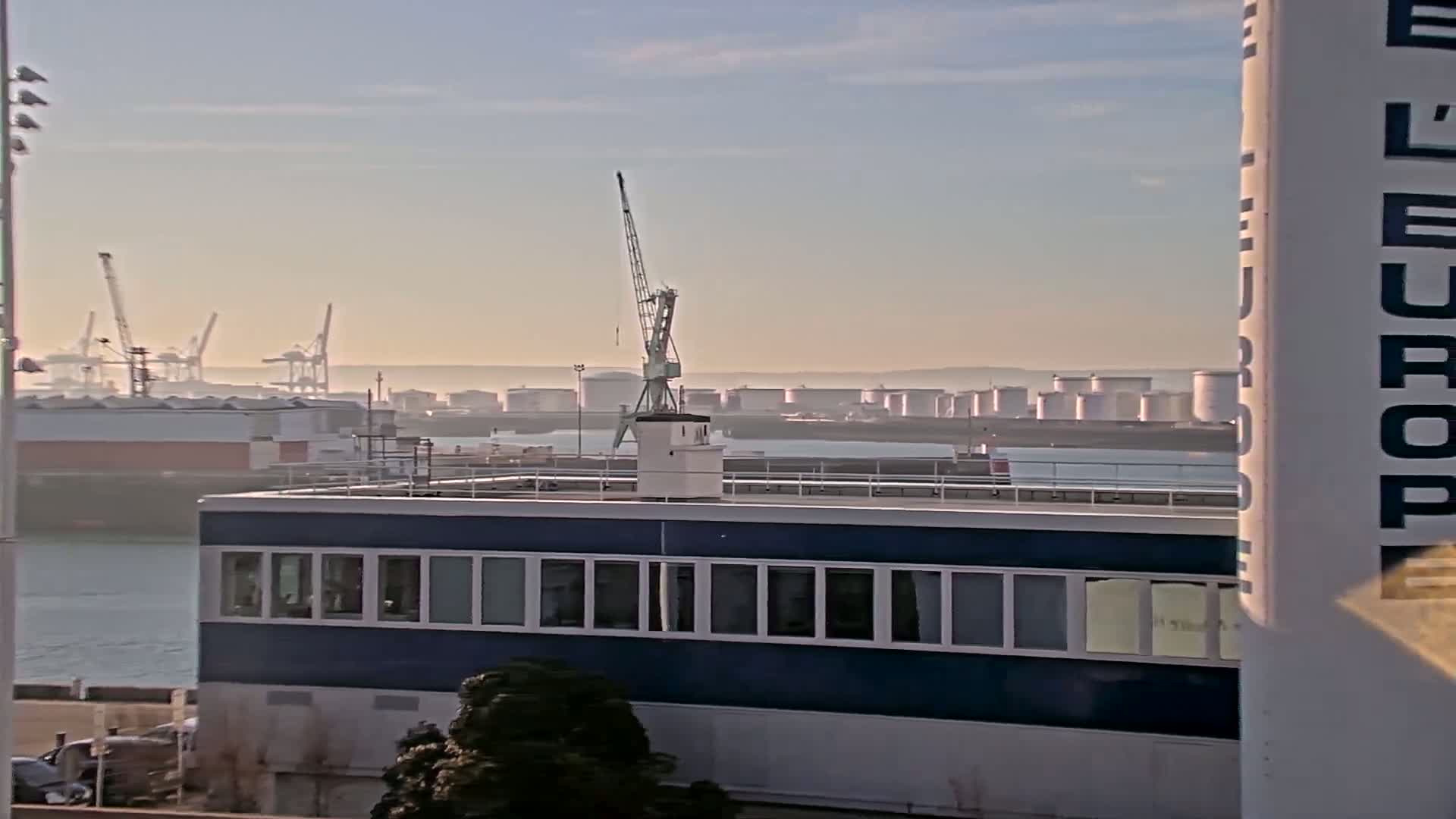A hazy sunrise illuminates a harbor scene featuring a blue building, a crane, numerous industrial storage tanks, and several cargo cranes in the distance.