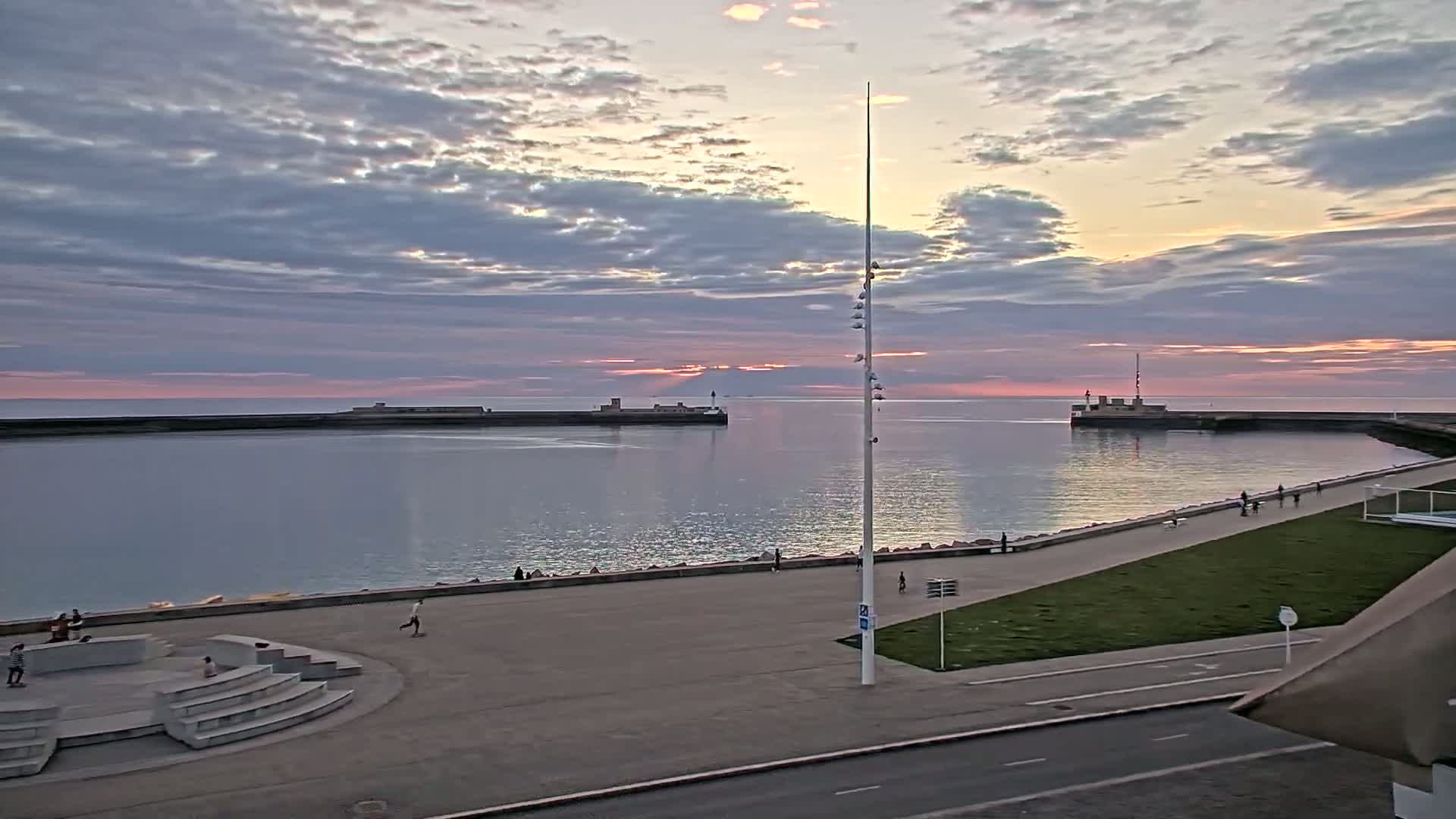 A calm harbor at sunset, with a paved walkway and a few people strolling along the shore under a partly cloudy sky.