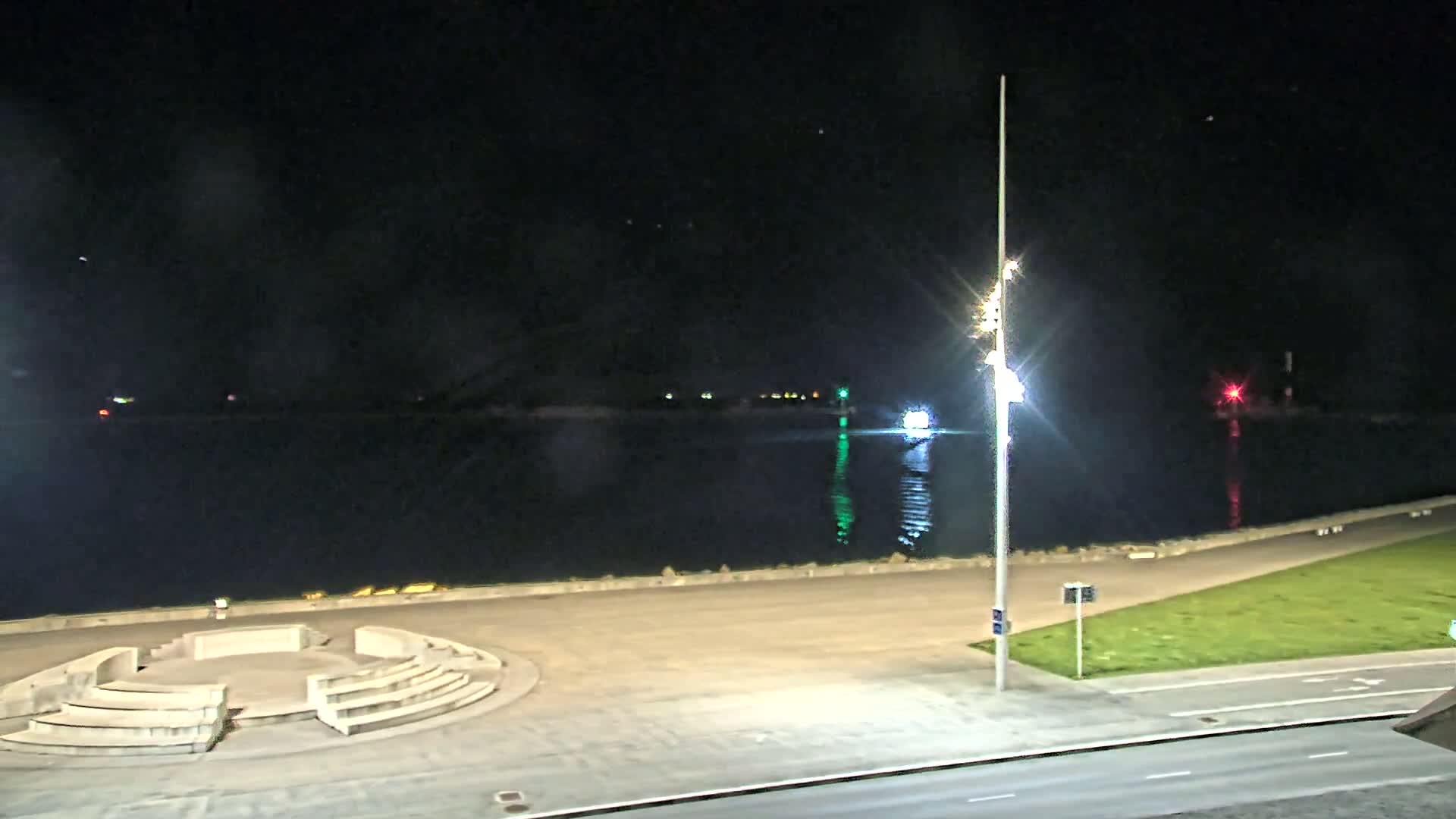 A nighttime view of a calm body of water, a paved waterfront area with a circular seating structure, and a street.