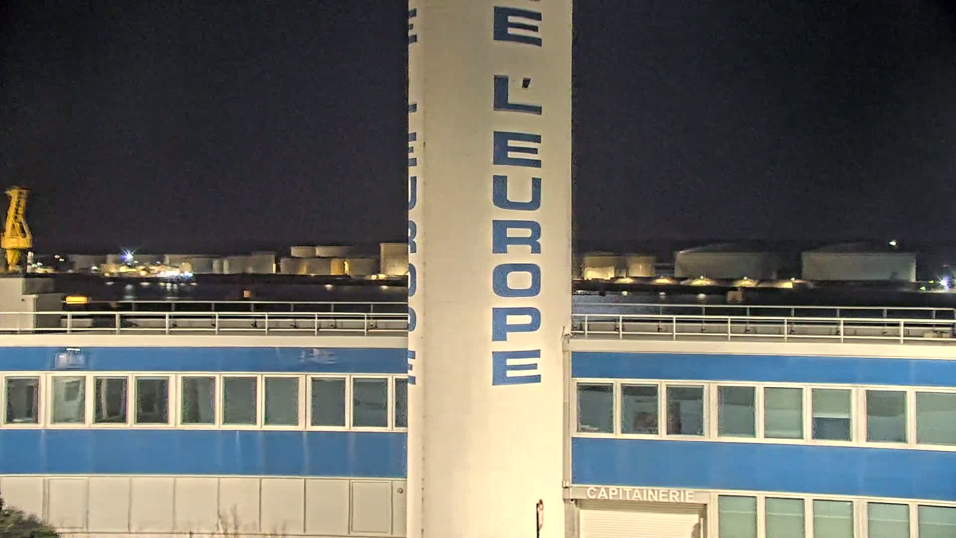 A tall, off-white structure with "L'EUROPE" written vertically in blue is seen next to a blue building at night, with a body of water and industrial structures visible in the background.