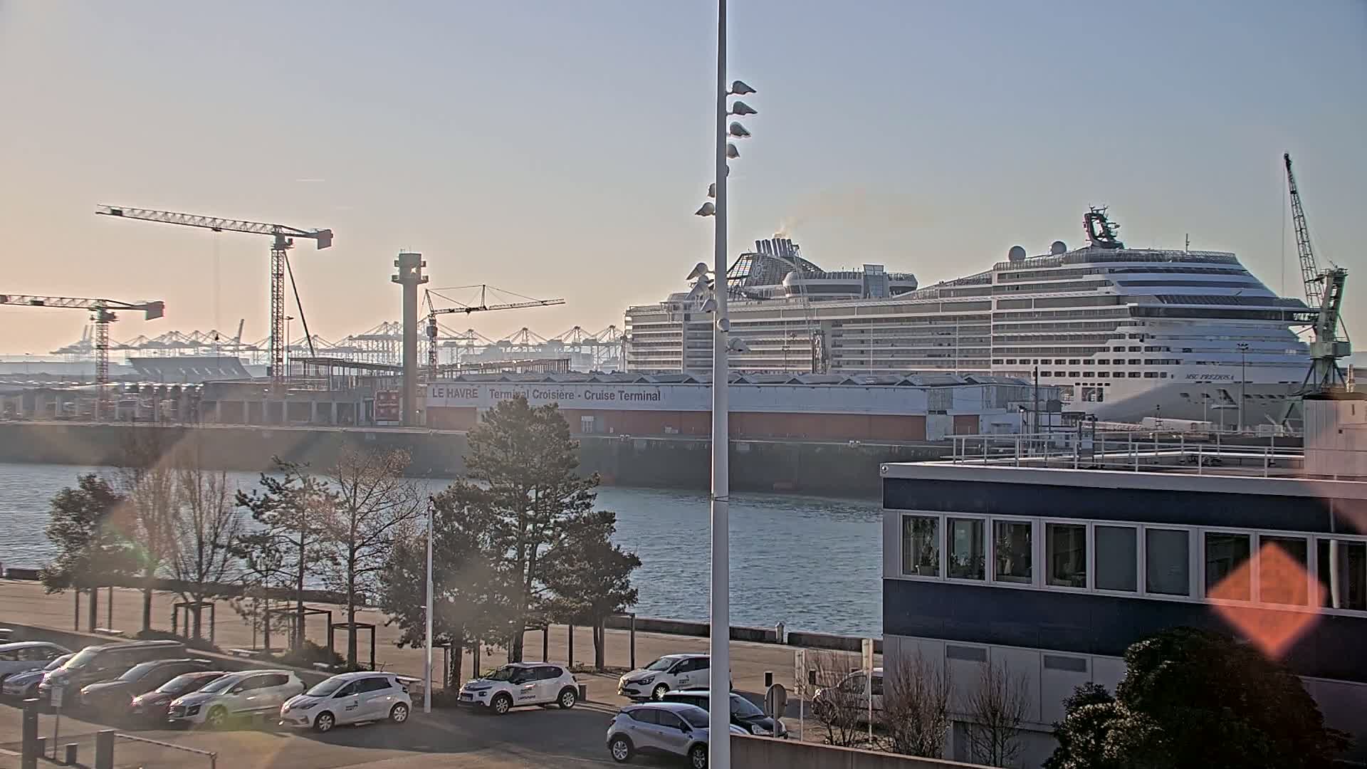 A large cruise ship is docked at a port, with construction cranes and other harbor infrastructure visible in the background under a clear, bright sky.