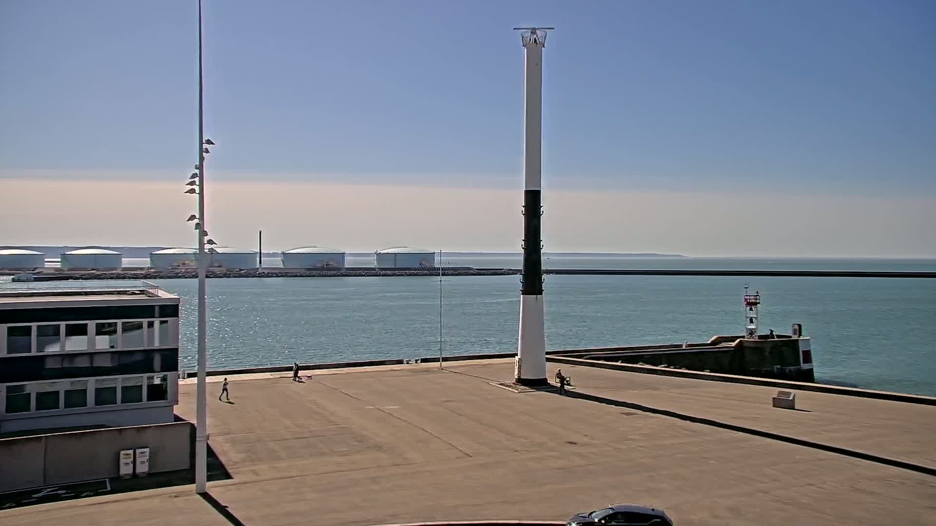 A tall, slender lighthouse stands in a harbor on a sunny day, with calm water, a distant breakwater, and several large storage tanks visible in the background.
