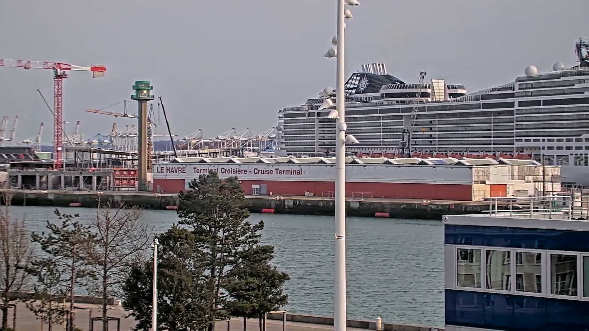 A large cruise ship is docked at a terminal next to a harbor with construction cranes and calm water under a mostly clear sky.