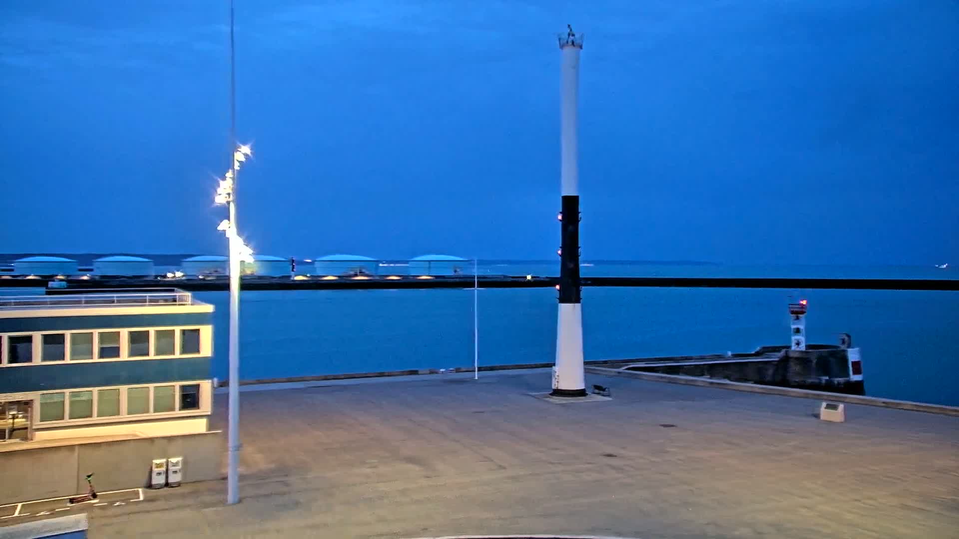 A harbor at twilight with a tall, slender white and black tower, a smaller lighthouse, and a building with blue siding, under a clear blue sky.