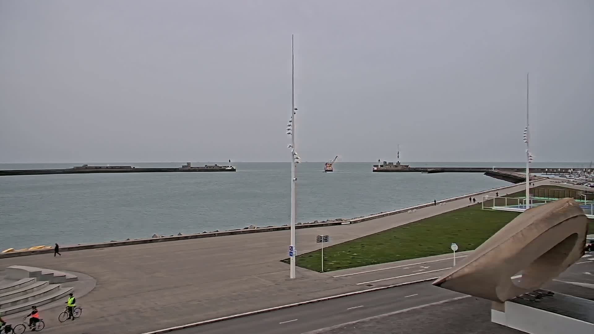 A harbor scene on an overcast day shows a calm body of water, breakwaters, a small boat, and a paved walkway with people walking and biking.