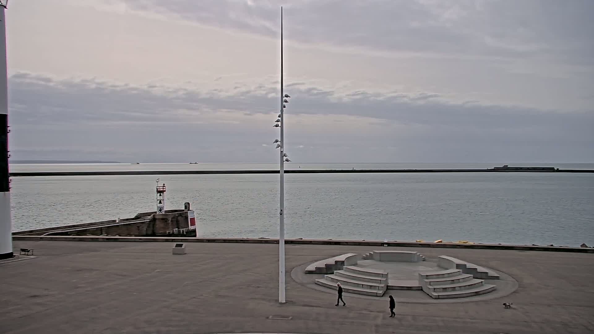 A harbor scene with overcast skies shows a paved area featuring a stone amphitheater, two pedestrians and a small dog, a lighthouse, and a tall slender pole with lights.