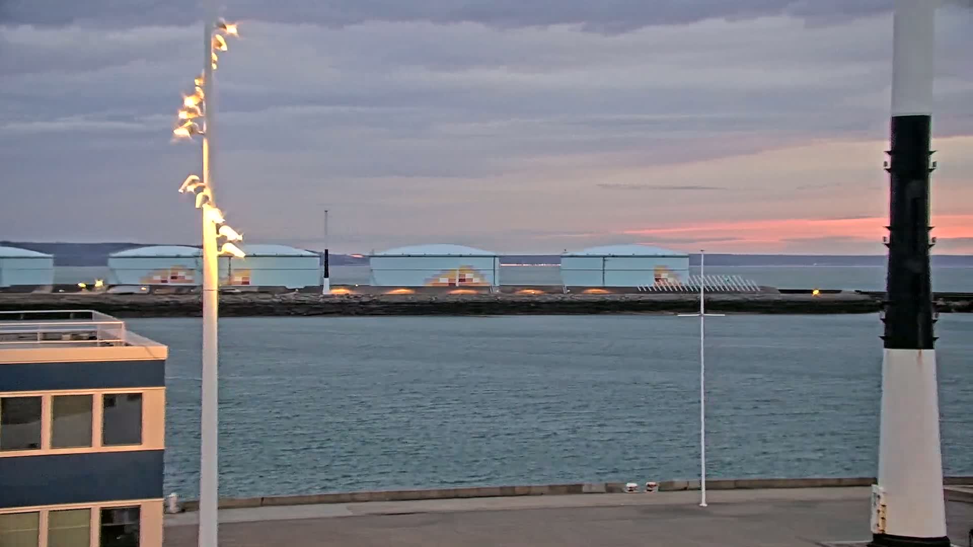 Several large, light-blue storage tanks sit on a rocky shoreline across a calm body of water under a dusky, pinkish-orange sky.