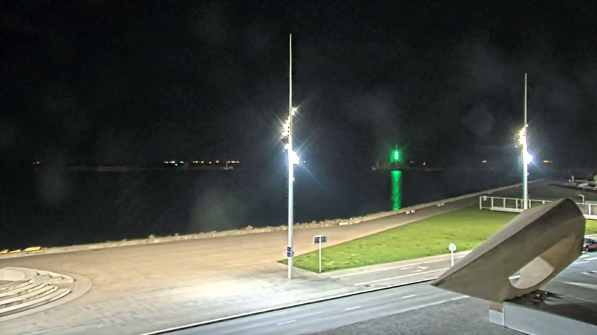 A nighttime view of a calm body of water next to a paved waterfront area with tall lights, a grassy area, and a roadway.