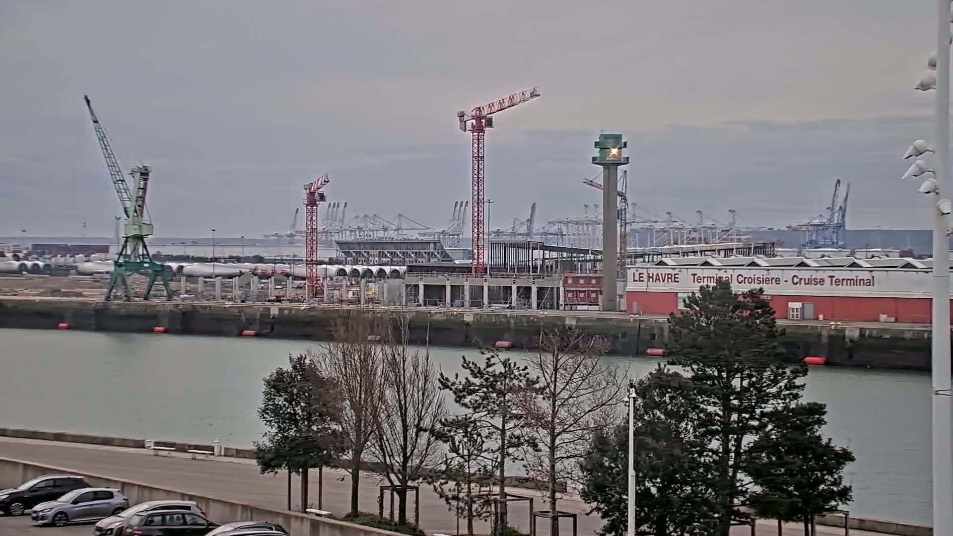 A harbor scene under an overcast sky shows a partially constructed cruise terminal, several cranes, and numerous container ships in the distance.