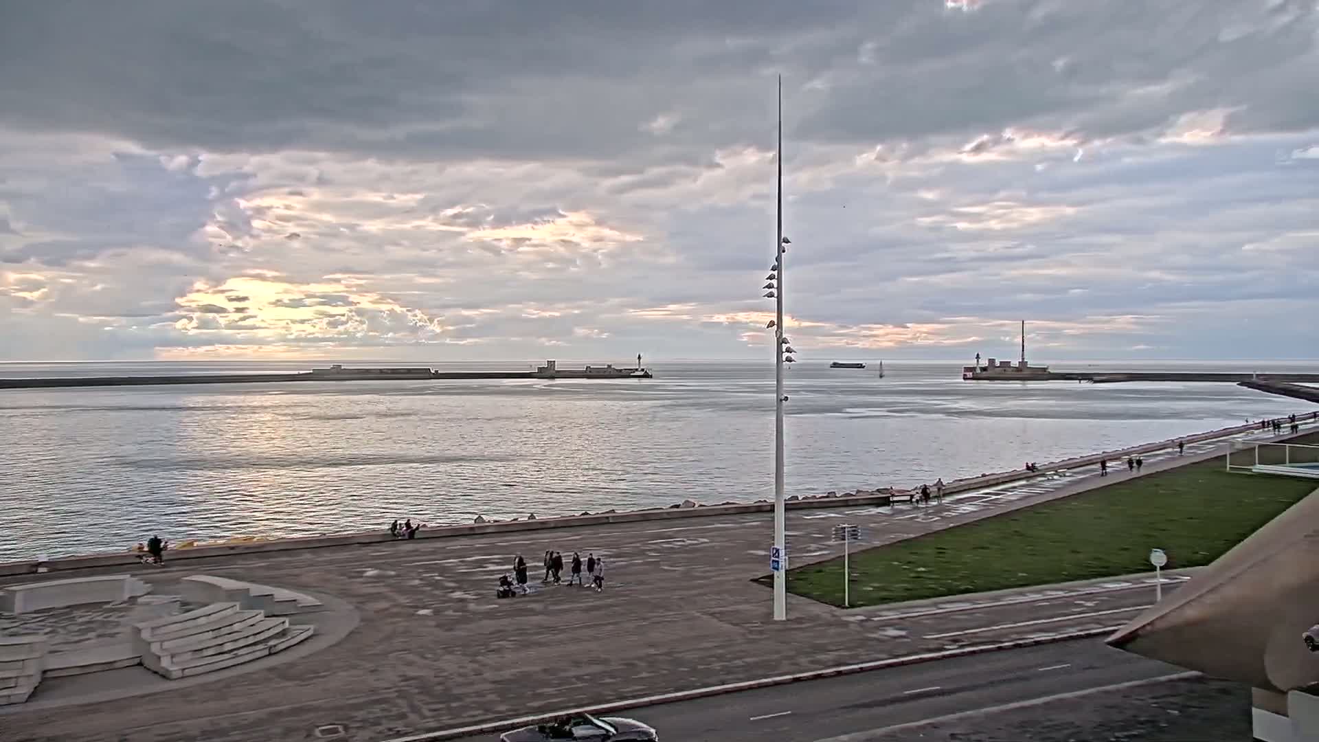 A calm body of water under a cloudy sky with some hints of sunset, is bordered by a paved walkway with people strolling along it and a grassy area, all overlooked by a tall light post.