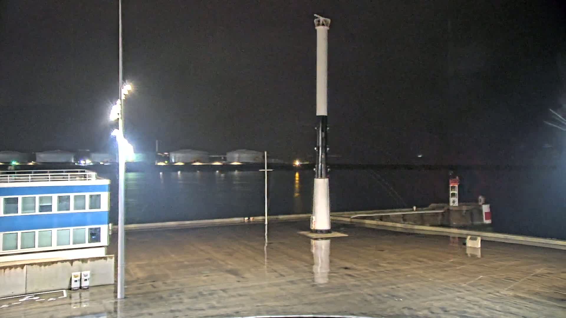 A tall, slender lighthouse stands on a wet, dark dock at night, with a body of water and industrial structures visible in the background under a dark sky.