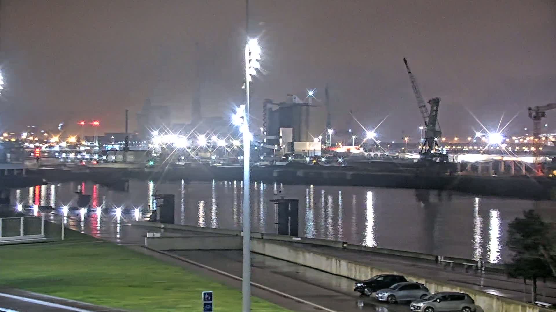 A nighttime view of a harbor with numerous lights reflecting on calm water, some cars parked nearby, and a crane visible in the distance under a hazy sky.