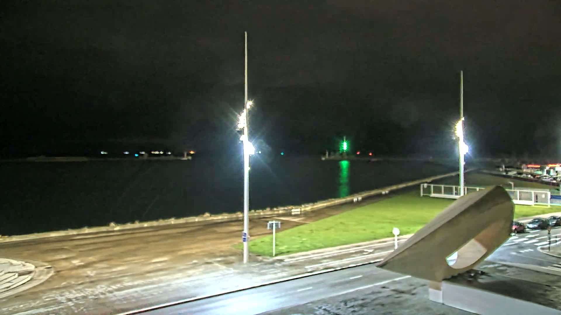 A nighttime view of a waterfront area with calm water, illuminated by tall lights, a grassy area, and a road, lightly dusted with snow.