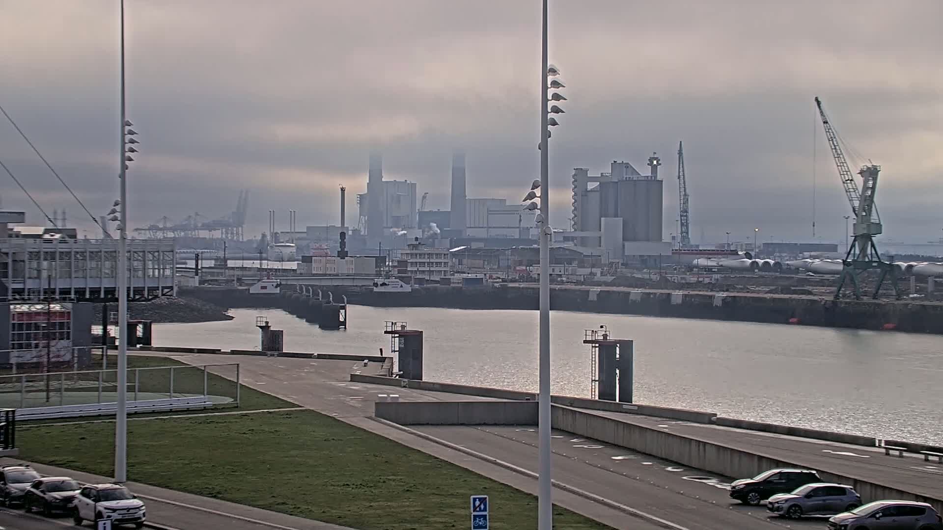 A foggy view of a harbor with industrial buildings in the background, a waterway in the foreground, and several cars parked along a road.