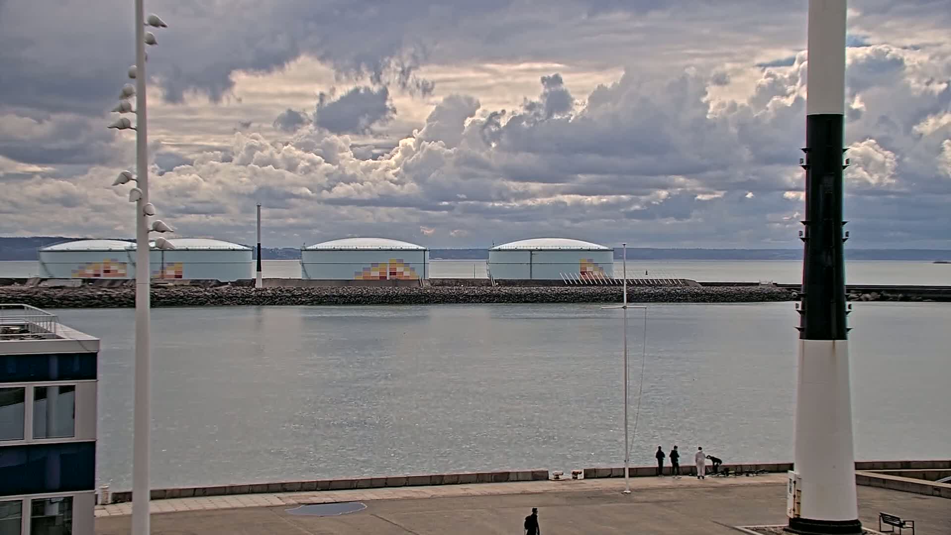 Four large, light-blue storage tanks with colorful geometric designs sit on a rocky jetty beside a calm body of water under a partly cloudy sky.