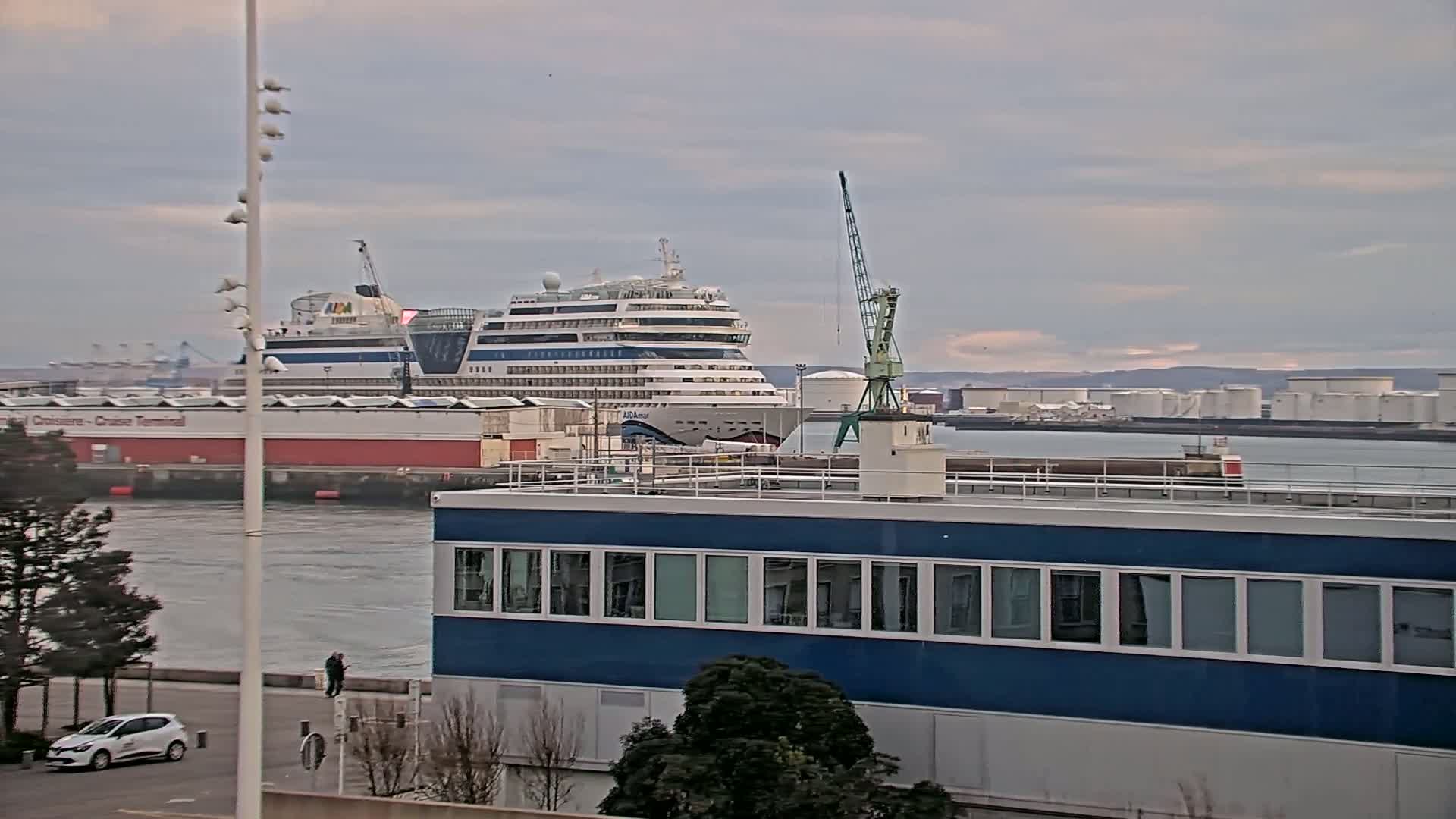 A large cruise ship is docked at a port, under a cloudy sky, near a building with many windows and a small crane.