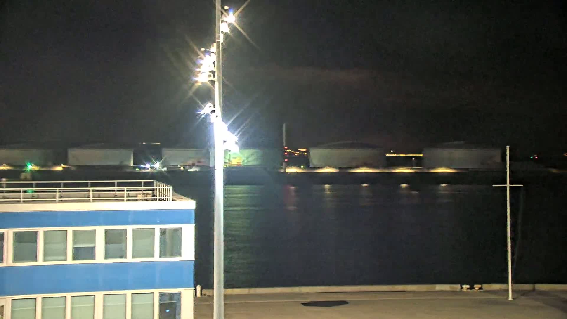 A brightly lit tall pole stands near a blue building overlooking dark water at night, with large storage tanks illuminated in the distance.