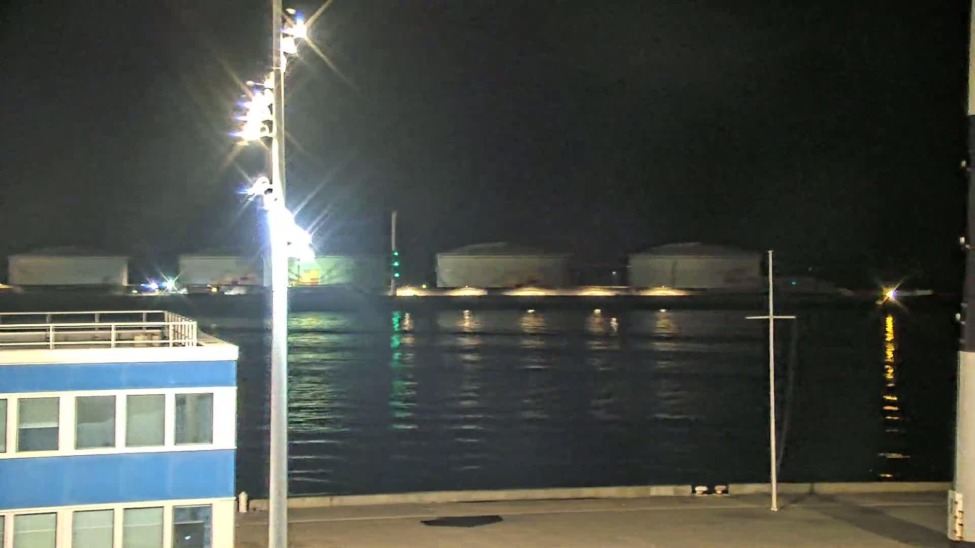 A nighttime view of a calm body of water reflecting lights from industrial structures across the water and a tall light post in the foreground near a blue building.