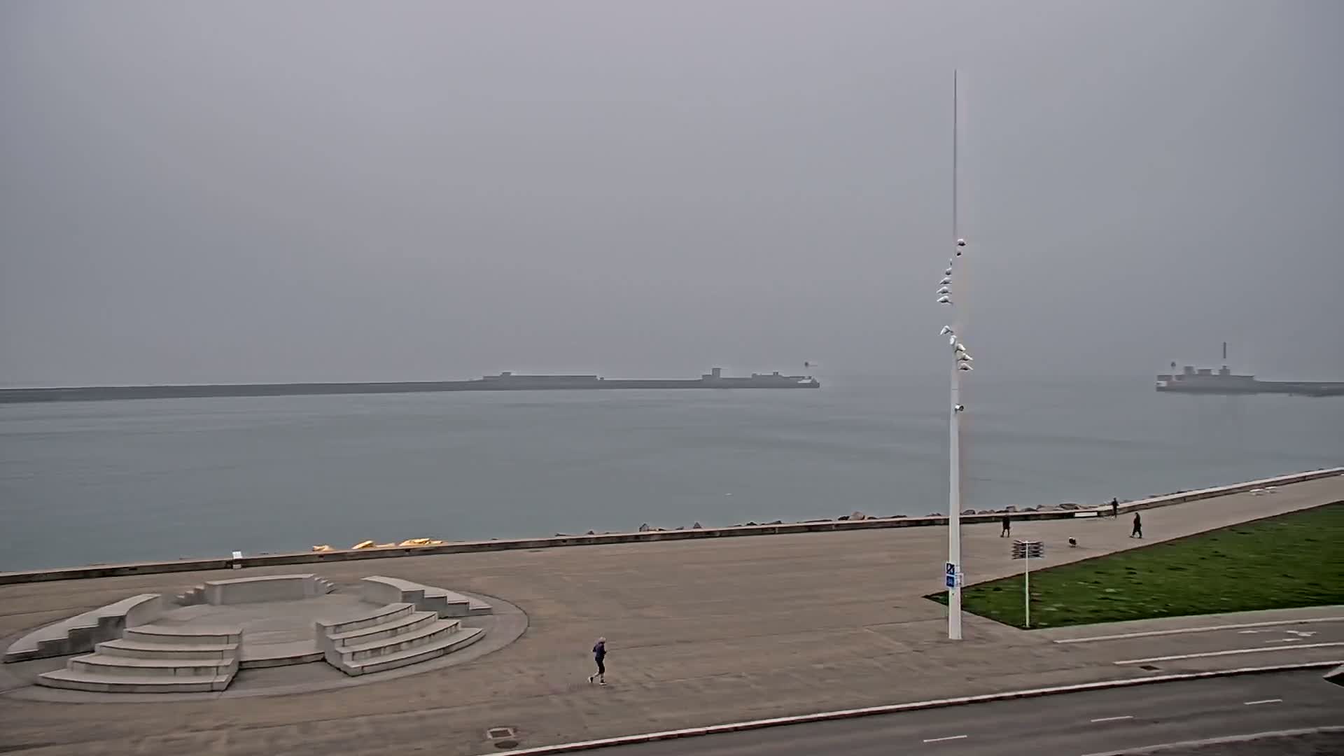 A mostly empty plaza overlooks a calm body of water on a foggy day, with a tall pole and a few people visible.