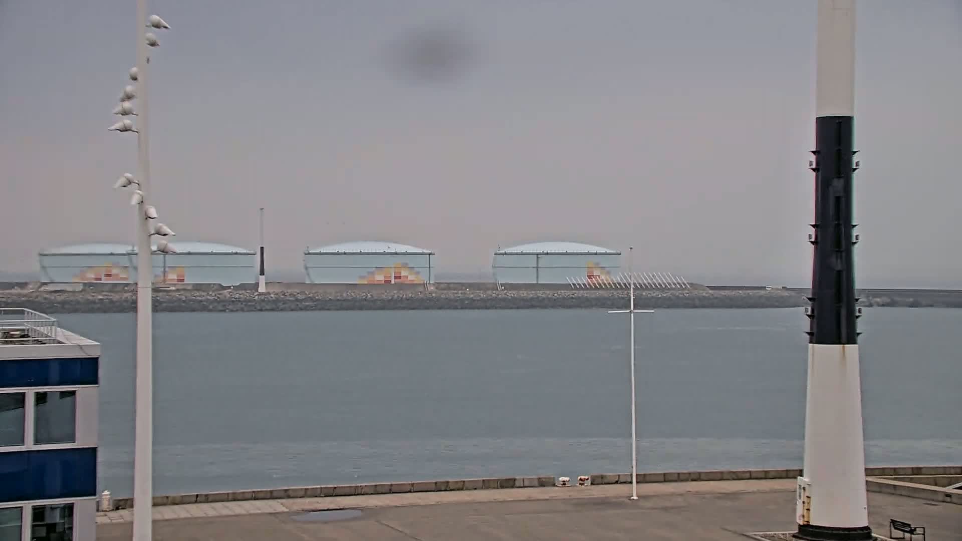 Three large, light-blue storage tanks sit on a rocky breakwater across a calm, gray body of water, under an overcast sky.