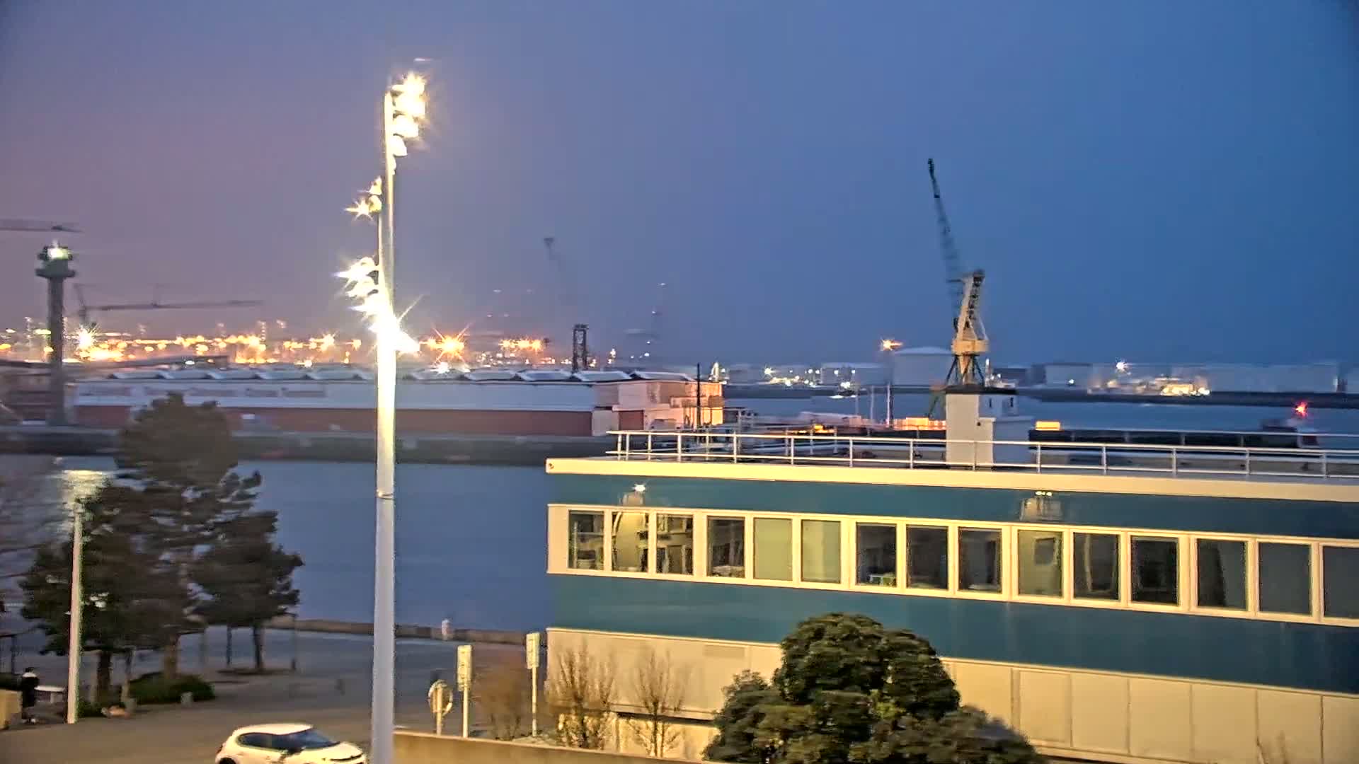 A nighttime view of a harbor with a teal building in the foreground, a body of water, ships, and industrial buildings across the water under a twilight sky.