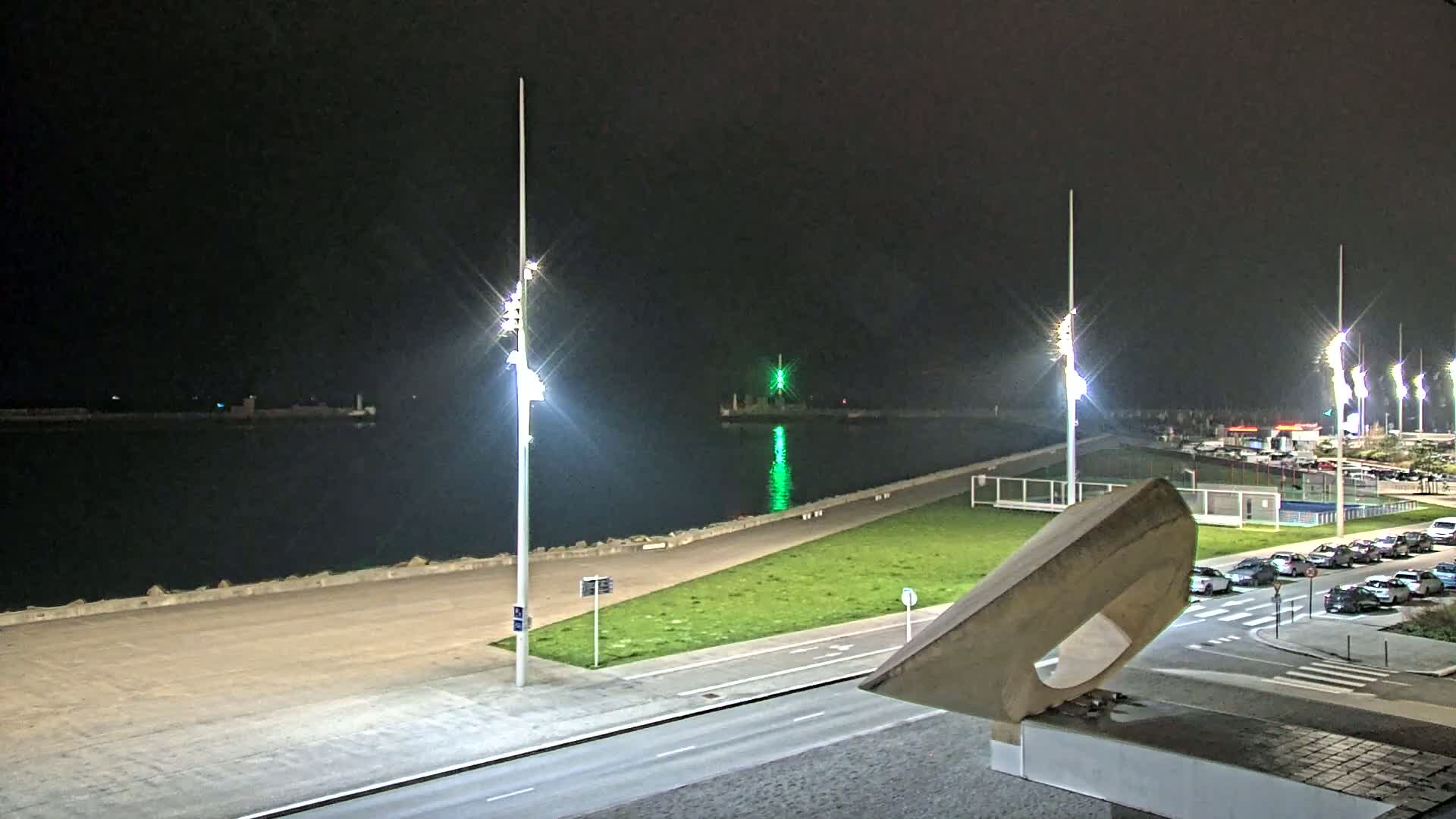 A nighttime view of a harbor with brightly lit walkways, a grassy area, parked cars, and tall light posts illuminating the scene.