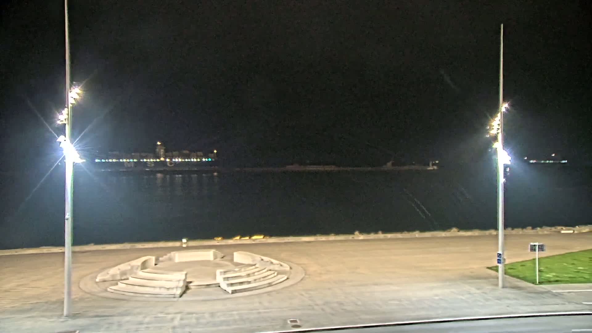 At night, a waterfront area with two tall light posts flanking a circular stone seating area overlooks a calm body of water with distant city lights across the water.